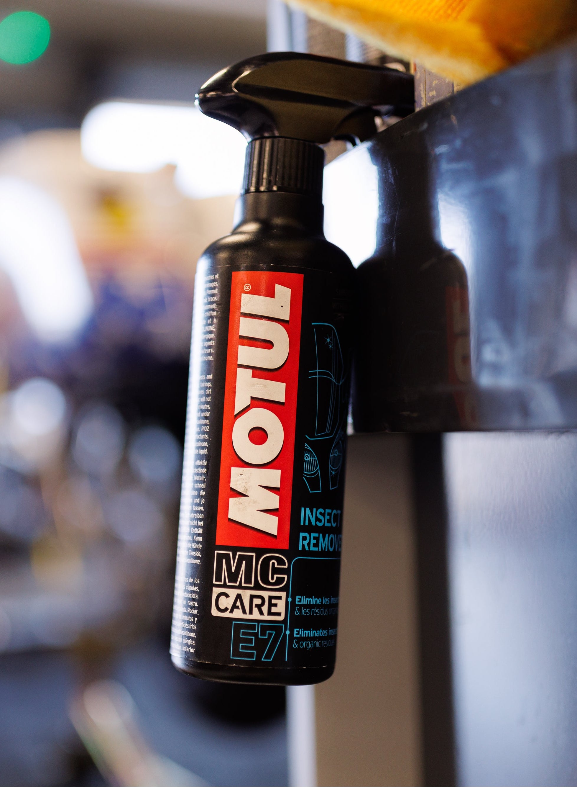 A bottle of Motul MC Care insect remover hanging on a dark metallic surface in a blurred workshop or garage setting.