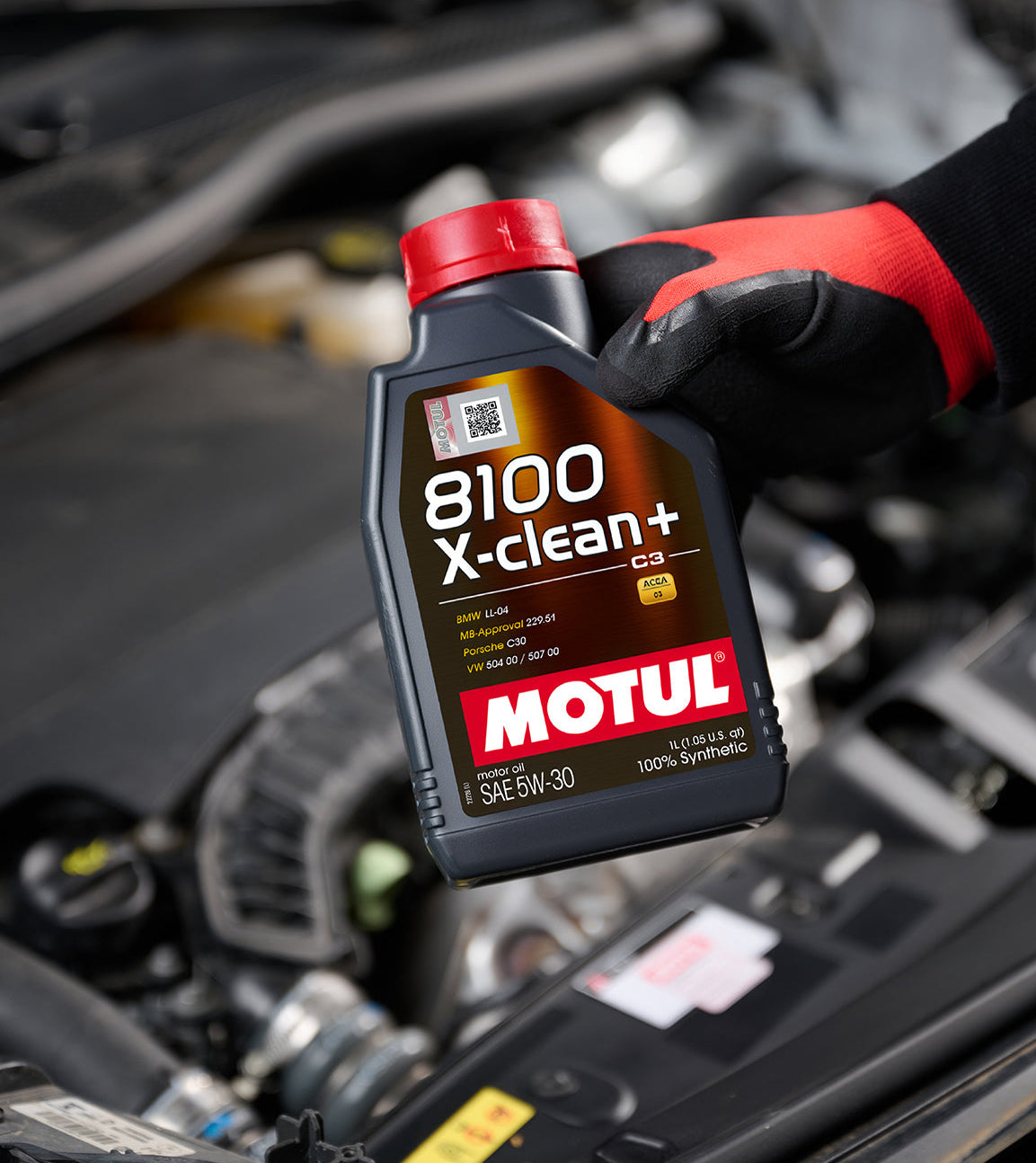 Hand holding a bottle of Motul 8100 X-clean+ oil in front of an open car hood.

