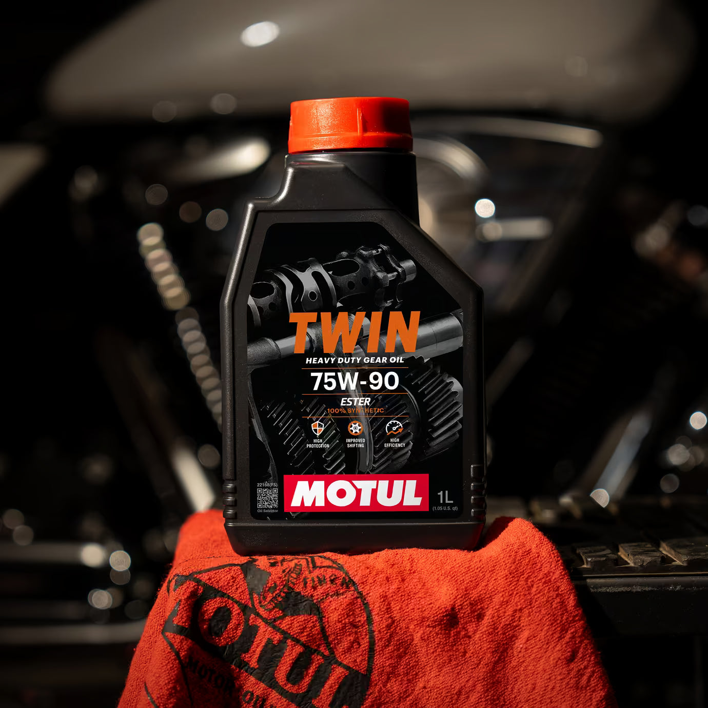 Motul Twin heavy duty gear oil bottle with red cap on an orange towel against a blurred car engine background