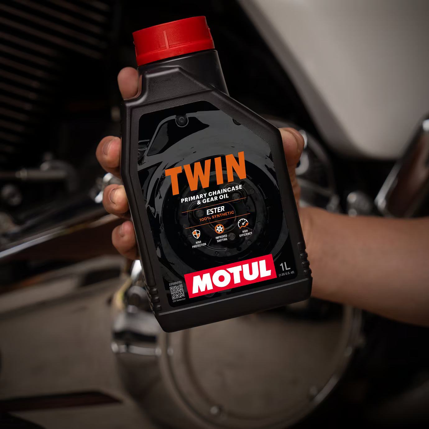 Hand holding a bottle of Motul Twin engine oil with a motorcycle in the background